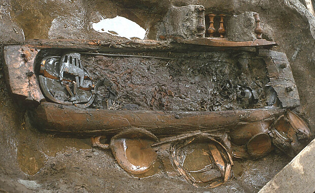 Eine feuchte Grube mit einem geöffnetem Sarg, man sieht viele Grabbeigaben aus Holz und eine Abdeckung aus Textil