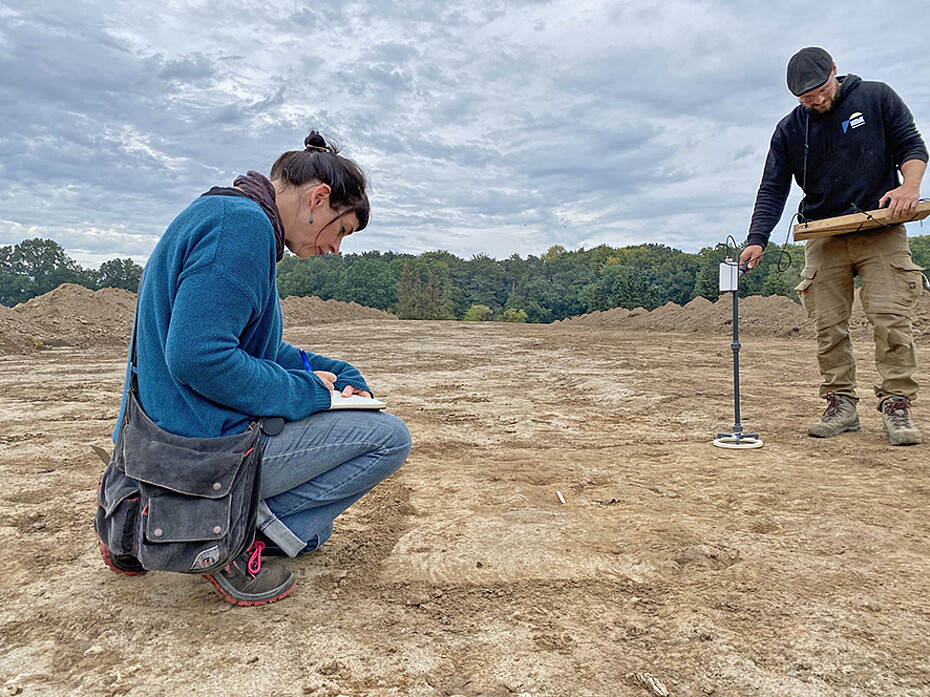 Archäologin und Techniker auf einer sandigen Fläche machen sich Notizen
